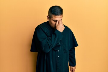Young latin priest man standing over yellow background tired rubbing nose and eyes feeling fatigue...