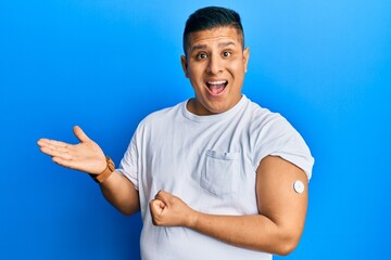 Young latin man using glucose meter button celebrating achievement with happy smile and winner expression with raised hand - obrazy, fototapety, plakaty