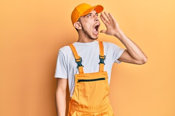 Hispanic young man wearing handyman uniform shouting and screaming loud to side with hand on mouth. communication concept.