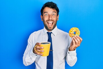 Handsome business man with beard eating doughnut and drinking coffee celebrating crazy and amazed for success with open eyes screaming excited.