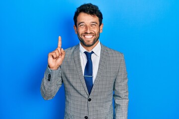 Handsome man with beard wearing business suit and tie showing and pointing up with finger number one while smiling confident and happy.