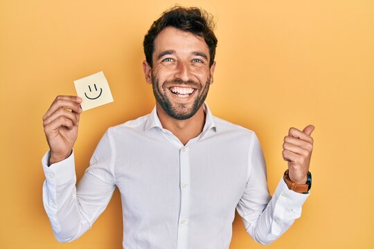 Handsome Man With Beard Holding Smile Emoji Reminder Pointing Thumb Up To The Side Smiling Happy With Open Mouth