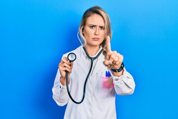 Beautiful young blonde doctor woman holding stethoscope pointing with finger up and angry expression, showing no gesture