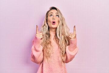 Beautiful young blonde woman wearing pink sweater amazed and surprised looking up and pointing with fingers and raised arms.