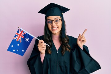 Young hispanic woman wearing graduation uniform holding australia flag smiling happy pointing with hand and finger to the side