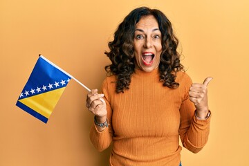 Middle age hispanic woman holding bosnia herzegovina flag pointing thumb up to the side smiling happy with open mouth