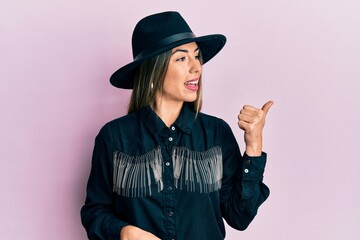 Fototapeta premium Young hispanic woman wearing cowgirl style smiling with happy face looking and pointing to the side with thumb up.