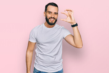 Fototapeta na wymiar Young man with beard wearing casual white t shirt smiling and confident gesturing with hand doing small size sign with fingers looking and the camera. measure concept.