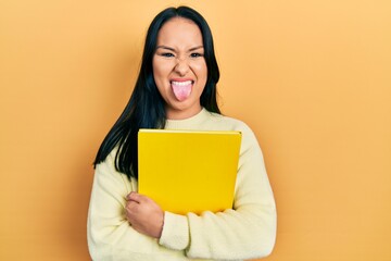 Beautiful hispanic woman with nose piercing holding book sticking tongue out happy with funny...