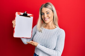 Beautiful blonde woman holding clipboard with blank space looking positive and happy standing and...