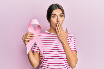 Young hispanic woman holding pink cancer ribbon covering mouth with hand, shocked and afraid for mistake. surprised expression