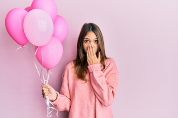 Young brunette woman holding pink balloons covering mouth with hand, shocked and afraid for...