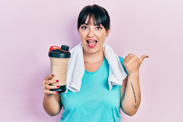 Young hispanic woman wearing sport clothes drinking a protein shake pointing thumb up to the side smiling happy with open mouth