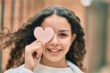 Hispanic teenager girl smiling happy holding heart over eye at the city.