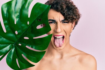 Young man wearing woman make up holding green plant leaf close to beautiful face sticking tongue...