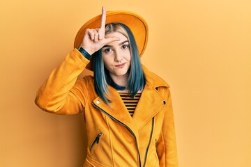 Young modern girl wearing yellow hat and leather jacket making fun of people with fingers on...