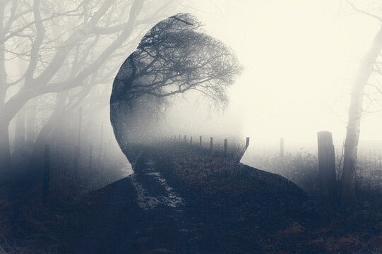 A double exposure of a spooky half transparent hooded figure. Over layered over a foggy path in the countryside. On a moody foggy winters day.