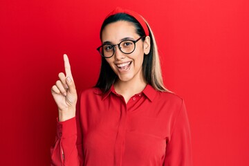 Young hispanic woman wearing casual clothes and glasses smiling with an idea or question pointing...