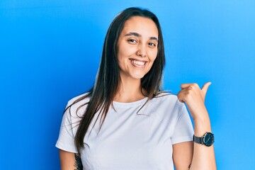 Young hispanic woman wearing casual white t shirt smiling with happy face looking and pointing to the side with thumb up.