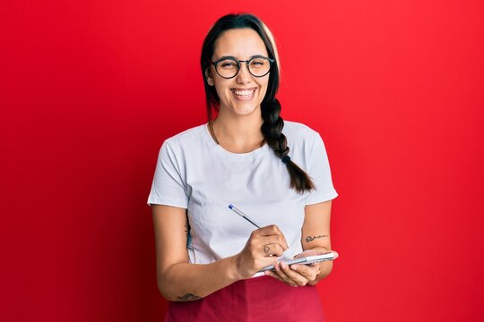 Young hispanic woman wearing waitress apron taking order smiling and laughing hard out loud because funny crazy joke.