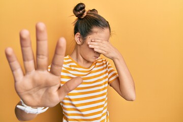 Beautiful brunette little girl wearing casual striped t shirt covering eyes with hands and doing...