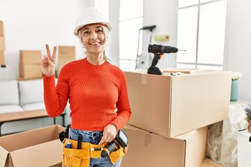 Middle age grey-haired woman wearing hardhat standing at new home showing and pointing up with fingers number two while smiling confident and happy.