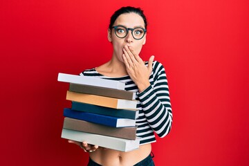 Young caucasian woman wearing glasses and holding books covering mouth with hand, shocked and afraid for mistake. surprised expression