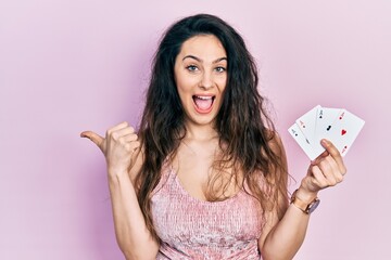 Young hispanic woman holding poker cards pointing thumb up to the side smiling happy with open mouth