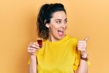 Young hispanic woman drinking whiskey shot pointing thumb up to the side smiling happy with open...