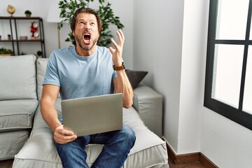 Handsome middle age man using computer laptop on the sofa crazy and mad shouting and yelling with...