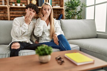 Young beautiful couple sitting on the sofa at home covering mouth with hand, shocked and afraid for mistake. surprised expression