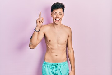 Fototapeta premium Young hispanic man wearing swimwear shirtless pointing finger up with successful idea. exited and happy. number one.