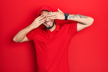 Hispanic man with beard wearing delivery uniform and cap covering eyes with hands smiling cheerful and funny. blind concept.