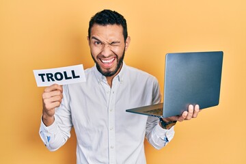 Hispanic man with beard working using computer laptop holding banner with troll word winking...