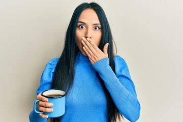 Young hispanic girl holding coffee covering mouth with hand, shocked and afraid for mistake....