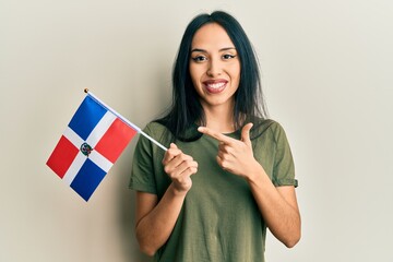 Young hispanic girl holding dominican republic flag smiling happy pointing with hand and finger - obrazy, fototapety, plakaty