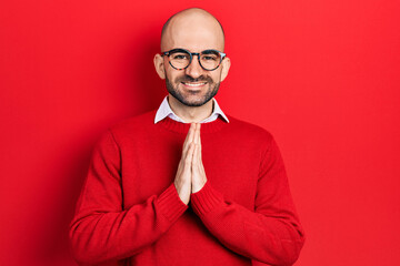 Young bald man wearing casual clothes and glasses praying with hands together asking for...