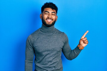 Fototapeta premium Arab man with beard wearing turtleneck sweater with a big smile on face, pointing with hand finger to the side looking at the camera.