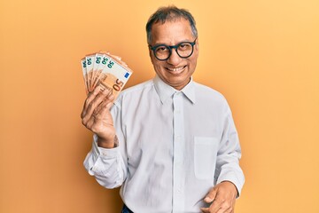 Middle age indian man holding bunch of 50 euro banknotes looking positive and happy standing and smiling with a confident smile showing teeth