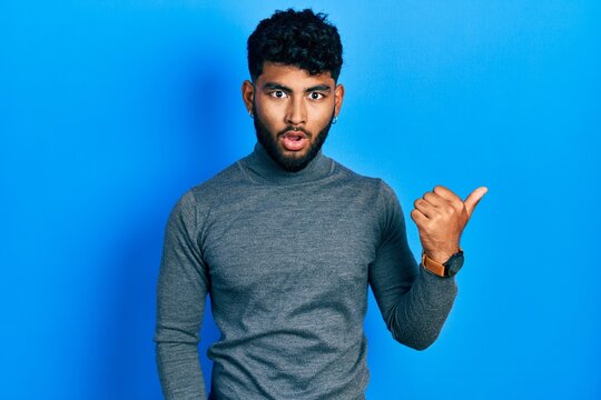 Arab man with beard wearing turtleneck sweater surprised pointing with hand finger to the side, open mouth amazed expression.