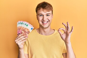 Young caucasian man holding bunch of 10 euro banknotes doing ok sign with fingers, smiling friendly gesturing excellent symbol