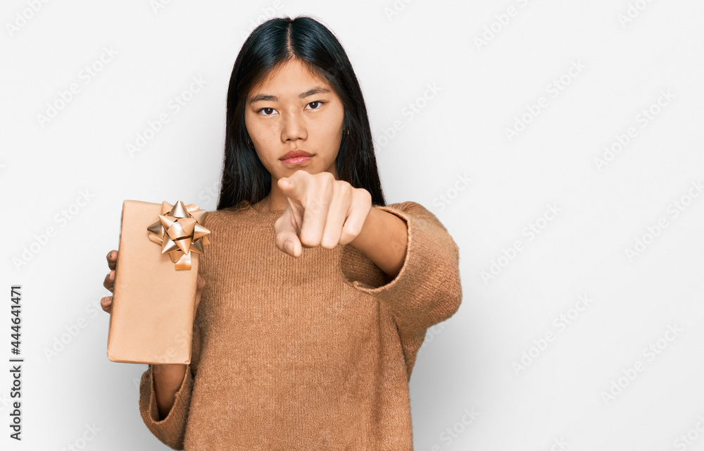 Poster Beautiful young asian woman holding gift pointing with finger to the camera and to you, confident gesture looking serious