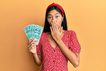 Young chinese woman holding 50 yuan chinese banknotes covering mouth with hand, shocked and afraid...