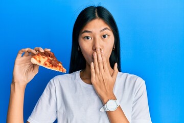 Young chinese woman eating tasty pepperoni pizza covering mouth with hand, shocked and afraid for...