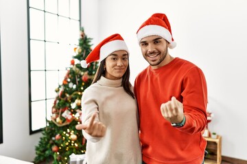 Young hispanic couple standing by christmas tree beckoning come here gesture with hand inviting welcoming happy and smiling