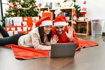 Young hispanic couple having video call using laptop lying on the floor by christmas tree at home.