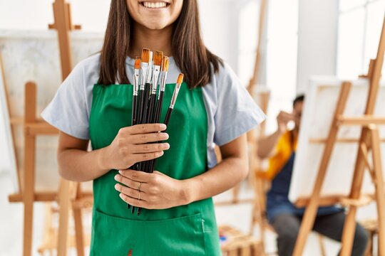 Young hispanic couple drawing at art studio. Woman smiling happy and holding paintbrushes.