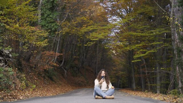Smiling long haired woman in loose sweater and jeans makes selfie sitting on empty asphalt road in picturesque autumn forest with old colorful trees