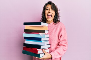 Young hispanic woman holding a pile of books celebrating crazy and amazed for success with open eyes screaming excited.