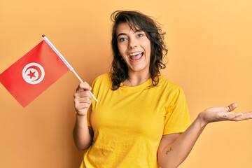 Young hispanic woman holding tunisia flag celebrating achievement with happy smile and winner expression with raised hand
